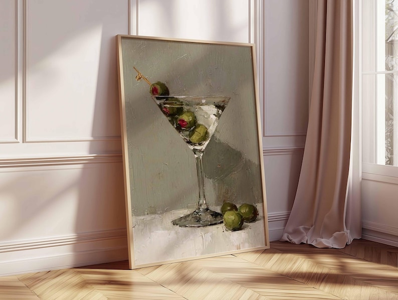 May include: A framed art print depicts a martini glass filled with clear liquid and green olives. The artwork is set against a muted green background, with the glass resting on a white surface. The print is displayed in a room with a wooden floor and a beige curtain.