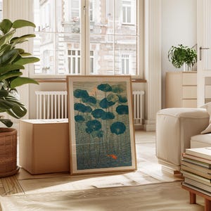 May include: A framed art print featuring a serene pond scene with teal lily pads and a small orange fish. The artwork has a vintage aesthetic, with a light brown wooden frame. The print is displayed in a well-lit living room setting.