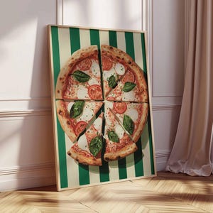 May include: A framed print of a pizza cut into slices, with tomatoes, mozzarella, and basil. The pizza is on a green and white striped background. The frame is leaning against a white wall and wooden floor.
