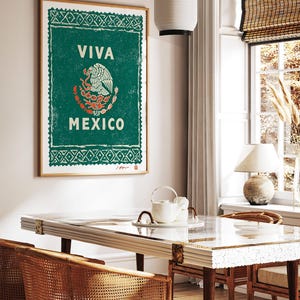 May include: A framed poster with a green background and the words "VIVA MEXICO" in white. The Mexican coat of arms is in the center. The poster is hanging on a wall above a table with woven chairs.