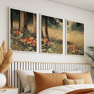 May include: Three framed art prints showing a forest scene with red and orange mushrooms. The artwork features tall trees, grassy areas, and a muted colour palette. The prints are displayed above a bed with orange and white pillows.