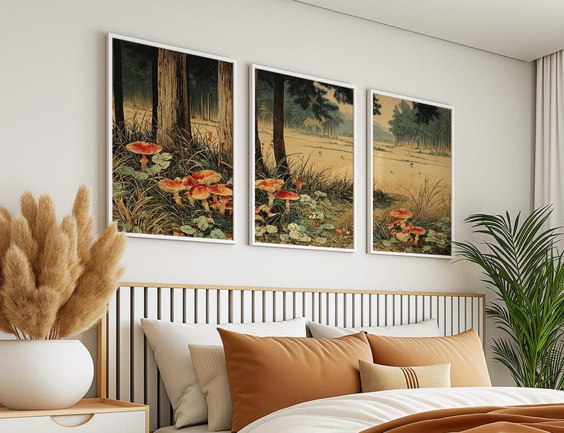 May include: Three framed art prints showing a forest scene with red and orange mushrooms. The artwork features tall trees, grassy areas, and a muted colour palette. The prints are displayed above a bed with orange and white pillows.