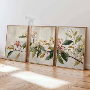 May include: Three framed botanical art prints. Each print features a branch with green leaves, small white flowers, and red and orange buds. The frames are a light brown color, and the background is a soft beige.