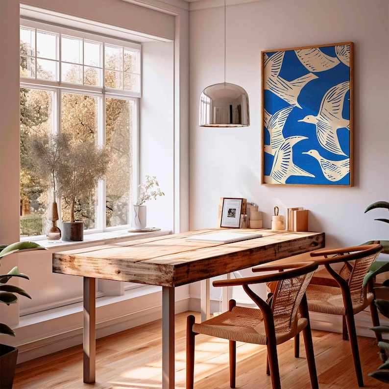 May include: A bright, airy room with a wooden desk and two chairs. A framed print with a blue background and white bird illustrations hangs on the wall. A large window offers a view of trees. A silver pendant light hangs above the desk.