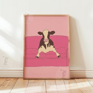May include: A framed art print showing a cow relaxing on a pink sofa. The cow is illustrated in black and white, with a cream-coloured body. The background is a gradient of pink shades, creating a whimsical and modern aesthetic.