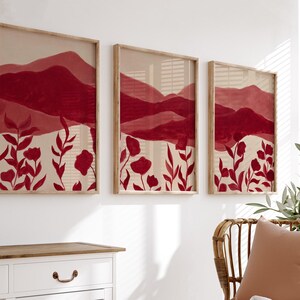 May include: Three framed art prints featuring a watercolor landscape with red mountains and floral elements. The artwork has a neutral background with red flowers and foliage. The frames are made of light-colored wood, creating a cohesive wall art set.