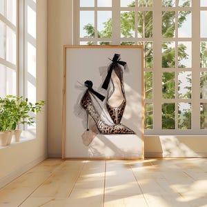 Op de afbeelding: Een ingelijste print van hoge hakken. Het kunstwerk toont twee elegante schoenen met spitse tenen en stilettohakken. E&eacute;n schoen heeft een luipaardprint, en beide hebben zwarte strikaccenten. De print wordt tentoongesteld in een kamer met natuurlijk licht.