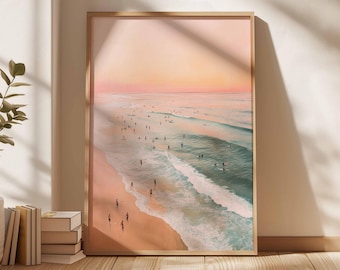 May include: A framed print of a beach scene at sunset. The artwork depicts a sandy beach with people, waves, and surfers. The sky transitions from pink to orange. The frame is a light wood color.