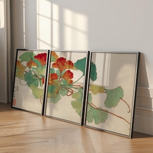May include: Three framed art prints featuring a botanical illustration of ginkgo leaves and flowers. The artwork showcases red and orange flowers with green and yellow leaves, set against a cream background. The frames are black with silver borders, leaning against a white wall.