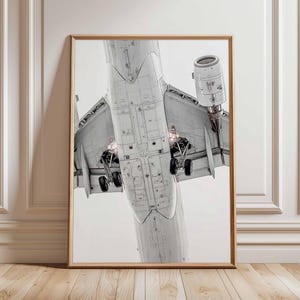 Könnte beinhalten: Gerahmter Druck eines Flugzeugs von unten, der Rumpf, Flügel und Fahrwerk zeigt. Das Bild ist in Graustufen gehalten und betont die technischen Details. Der Druck befindet sich in einem Holzrahmen.