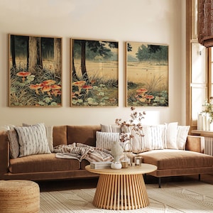 May include: Three framed art prints showing a forest scene with mushrooms. The artwork uses a muted colour palette of greens, browns, and reds. The prints are displayed above a brown sofa with decorative cushions, a round wooden coffee table, and a woven rug.