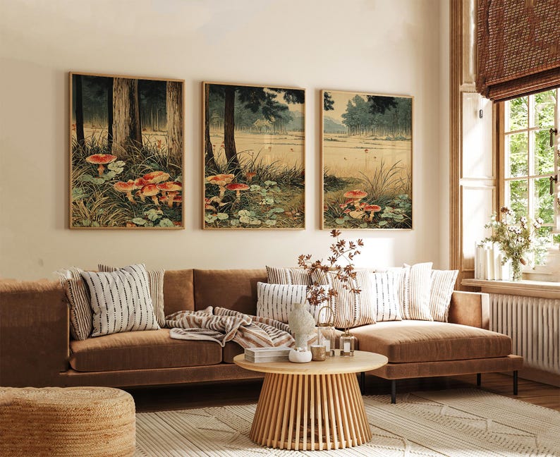 May include: Three framed art prints showing a forest scene with mushrooms. The artwork uses a muted colour palette of greens, browns, and reds. The prints are displayed above a brown sofa with decorative cushions, a round wooden coffee table, and a woven rug.