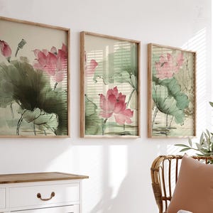 May include: Three framed art prints featuring watercolor paintings of pink lotus flowers and green leaves. The artwork has a vintage aesthetic, with a light beige background and wooden frames. The prints are displayed above a white dresser.