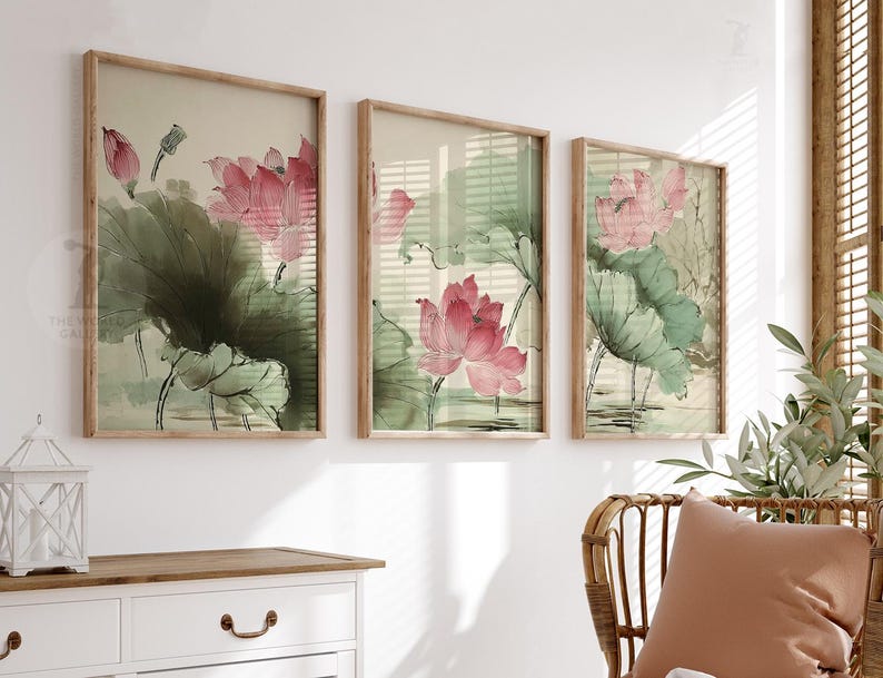 May include: Three framed art prints featuring watercolor paintings of pink lotus flowers and green leaves. The artwork has a vintage aesthetic, with a light beige background and wooden frames. The prints are displayed above a white dresser.