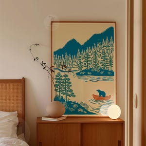 May include: A framed art print featuring a teal and cream illustration of a lake scene with mountains, trees, and a bear in a canoe. The artwork is displayed on a wooden cabinet with a round lamp and a decorative vase.