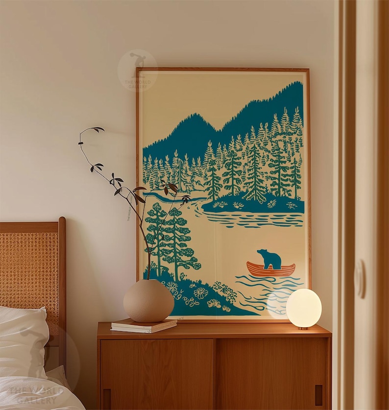 May include: A framed art print featuring a teal and cream illustration of a lake scene with mountains, trees, and a bear in a canoe. The artwork is displayed on a wooden cabinet with a round lamp and a decorative vase.