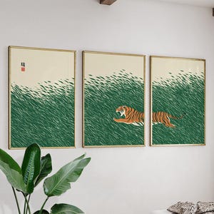 Peut inclure: Ensemble de trois impressions d'art encadrées. Chaque impression représente un tigre dans un champ d'herbe verte. Le tigre est orange et noir. Le fond est de couleur beige clair. Les cadres sont dorés.