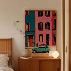 May include: A framed art print depicts a colorful European street scene with a teal car. The buildings are painted in pink and teal with black window shutters. A round, beige vase and a small lamp sit on a wooden cabinet.