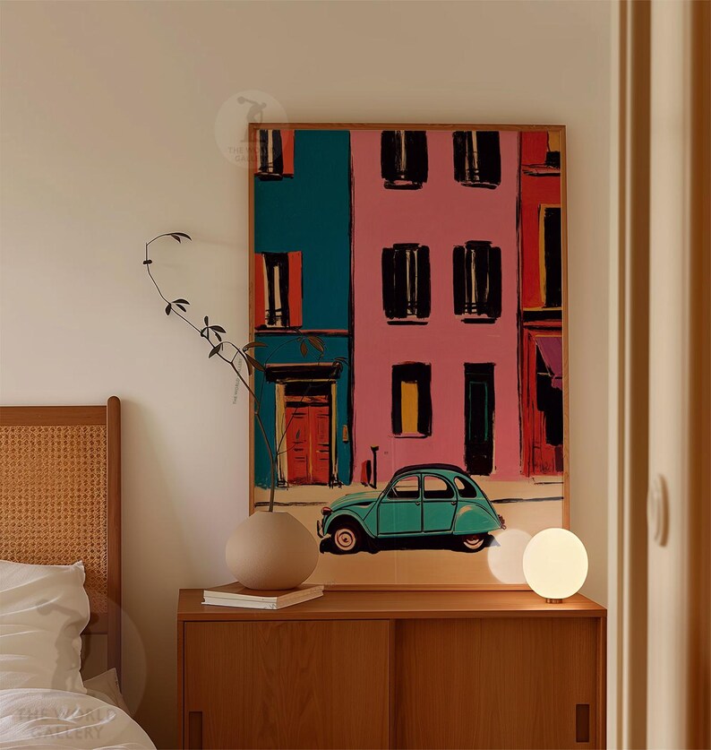 May include: A framed art print depicts a colorful European street scene with a teal car. The buildings are painted in pink and teal with black window shutters. A round, beige vase and a small lamp sit on a wooden cabinet.