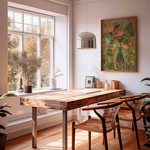 May include: A bright, airy room with a wooden desk and two chairs. A framed painting of a woman in a garden hangs on the wall. A large window offers a view of trees. A silver pendant light hangs above the desk.