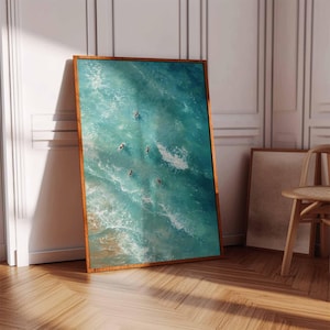 May include: A framed art print featuring an aerial view of turquoise ocean waves with several figures swimming. The artwork is displayed in a room with a wooden floor and a white wall, and is framed in a warm brown wood.