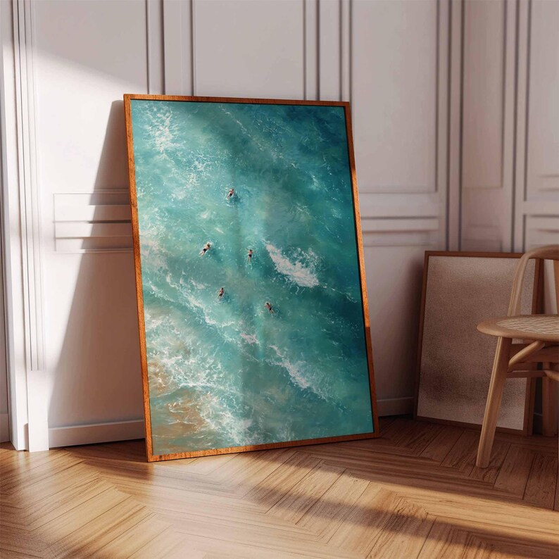 May include: A framed art print featuring an aerial view of turquoise ocean waves with several figures swimming. The artwork is displayed in a room with a wooden floor and a white wall, and is framed in a warm brown wood.