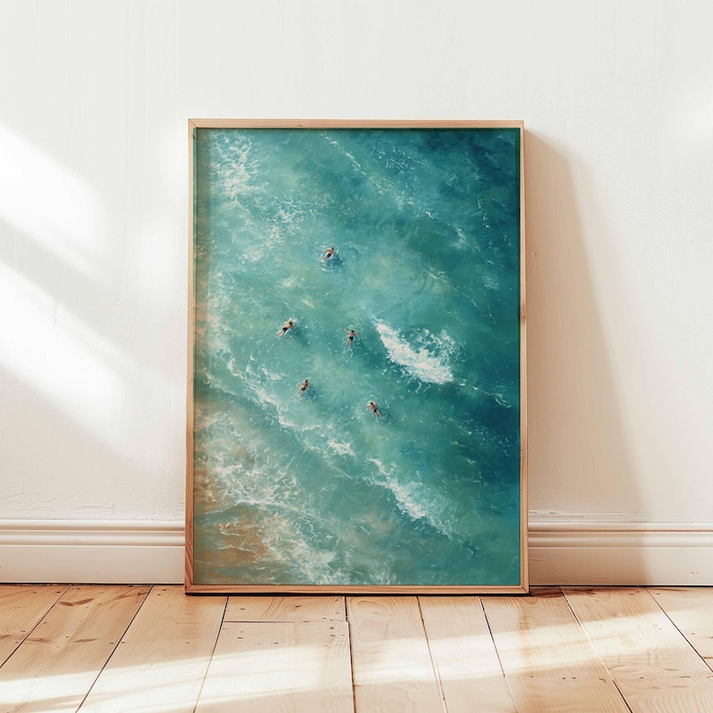 May include: A framed art print featuring an aerial view of swimmers in turquoise ocean water. The artwork captures the movement of the waves and the figures of the swimmers. The frame is a light brown color, and the artwork is displayed on a wooden floor.