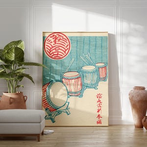 May include: A framed Japanese-style print featuring a red and blue illustration of drums and a red sun. The artwork is displayed in a room with a plant, a gray ottoman, and decorative pottery. The print has Japanese characters.