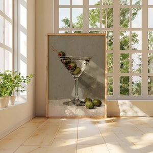 May include: A framed print of a martini glass filled with olives and a clear liquid. The artwork is displayed in a bright room with a window and a view of trees. The painting has a neutral color palette.