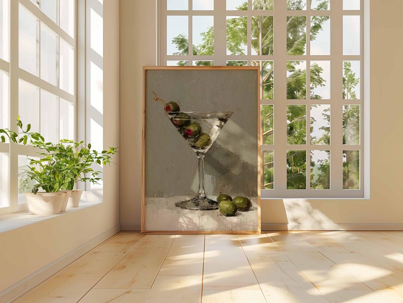 May include: A framed print of a martini glass filled with olives and a clear liquid. The artwork is displayed in a bright room with a window and a view of trees. The painting has a neutral color palette.