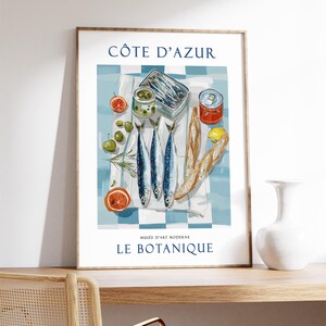May include: A framed art print featuring a still life of sardines, olives, and citrus fruits on a checkered tablecloth. The print includes the text "C&Ocirc;TE D'AZUR" and "MUS&Eacute;E D'ART MODERNE LE BOTANIQUE". The color palette includes blue, white, and orange.