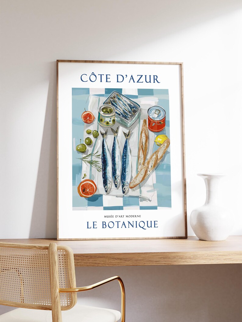 May include: A framed art print featuring a still life of sardines, olives, and citrus fruits on a checkered tablecloth. The print includes the text "C&Ocirc;TE D'AZUR" and "MUS&Eacute;E D'ART MODERNE LE BOTANIQUE". The color palette includes blue, white, and orange.