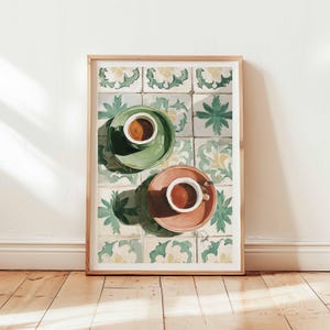 May include: Framed art print featuring two cups of coffee on saucers, one green and one peach, set against a backdrop of patterned tiles. The artwork is displayed in a light wood frame, leaning against a wall and wooden floor.