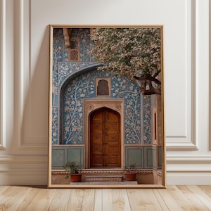 May include: Framed print of an ornate doorway with a wooden door, set against a teal wall adorned with floral patterns. A tree with white blossoms partially frames the entrance. Two potted plants sit at the base of the door.