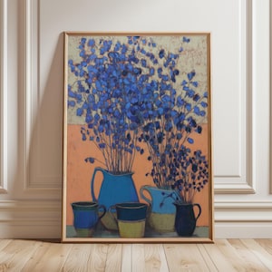 May include: A framed art print featuring a still life composition. The artwork depicts blue flowers in blue pitchers and mugs against a peach and beige background. The artwork is displayed on a wooden floor.