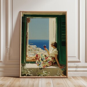 May include: A framed art print depicting a person sitting in an open window with green shutters, holding a glass of red wine. The person wears a white top and a floral skirt, overlooking a coastal town with white buildings and a blue sea.