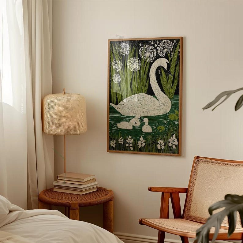 May include: A framed art print featuring a swan and two cygnets swimming in a pond. The artwork has a green, white, and black color scheme, with tall green reeds and white flowers in the background. The print is displayed on a white wall.