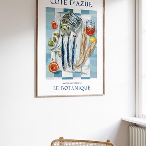 May include: A framed art print featuring a still life of French cuisine. The image includes sardines, olives, tomatoes, and bread on a blue and white checkered tablecloth. Text at the top reads "C&Ocirc;TE D'AZUR" and at the bottom "LE BOTANIQUE".