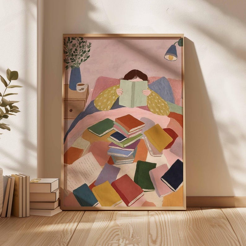 May include: A framed art print depicts a person reading in bed, surrounded by colorful books. The illustration features a warm color palette with a pink background, a wooden frame, and a cozy, inviting atmosphere.