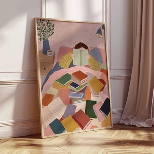 May include: A framed art print depicts a person reading in bed, surrounded by colorful books. The artwork features a soft color palette with pink, blue, and yellow tones. A vase with greenery and a bedside table are also visible.