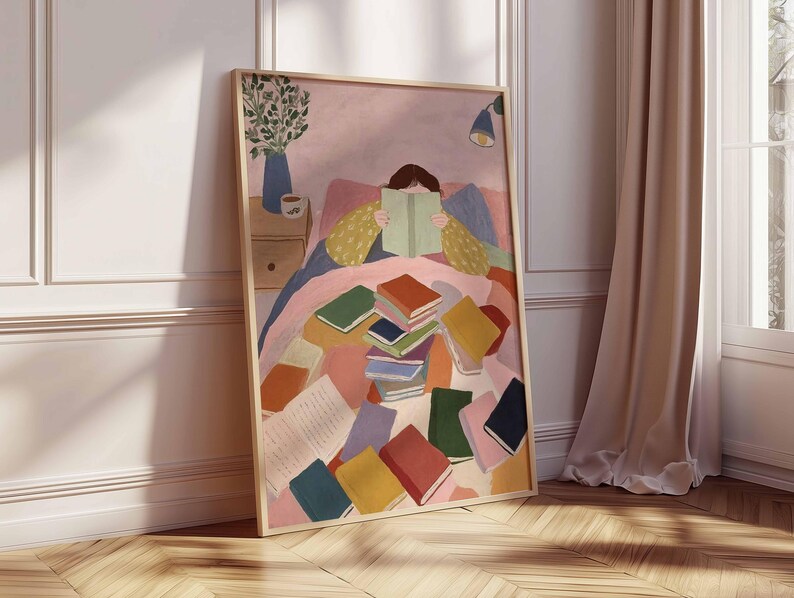 May include: A framed art print depicts a person reading in bed, surrounded by colorful books. The artwork features a soft color palette with pink, blue, and yellow tones. A vase with greenery and a bedside table are also visible.