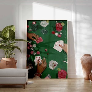May include: A framed art print depicts a poker game scene on a green felt table. Hands with rings hold playing cards, chips, and cocktails. The artwork is displayed in a room with a plant, ottoman, and decorative vases.