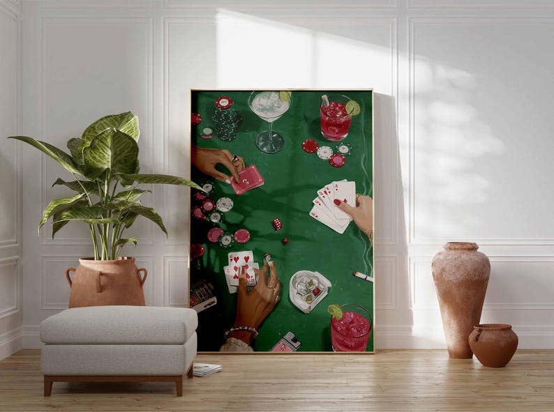 May include: A framed art print depicts a poker game scene on a green felt table. Hands with rings hold playing cards, chips, and cocktails. The artwork is displayed in a room with a plant, ottoman, and decorative vases.
