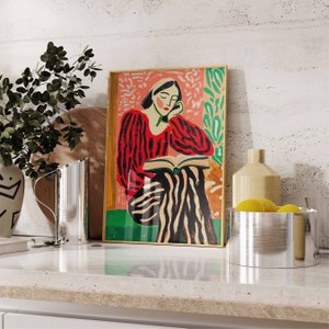 May include: A framed illustration of a woman in a red and black striped shirt reading a book. The woman is sitting on a striped chair with a pink background. The illustration is surrounded by a gold frame.