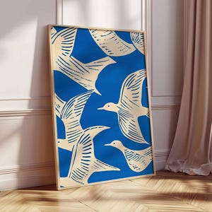 May include: A framed art print featuring a blue background with cream-colored birds in flight. The birds are stylized with detailed wing patterns. The artwork is displayed in a light-colored wooden frame, leaning against a white wall and wooden floor.
