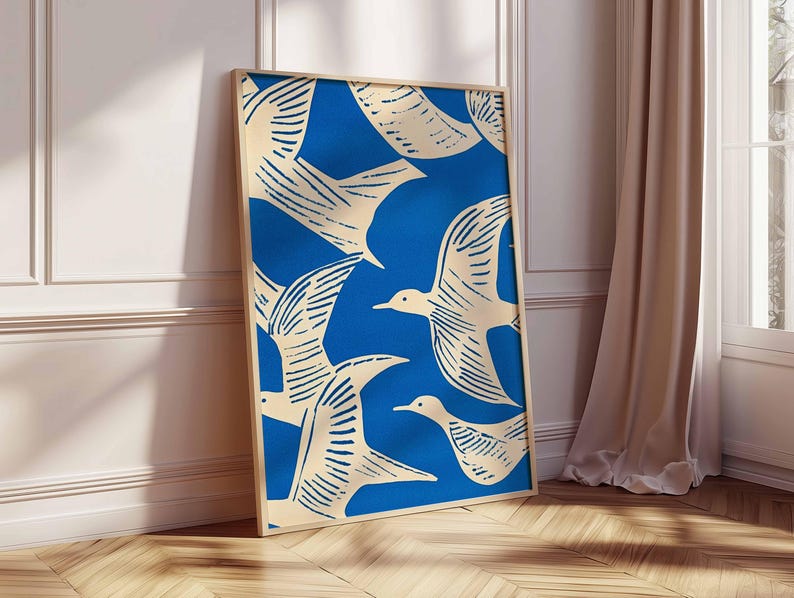 May include: A framed art print featuring a blue background with cream-colored birds in flight. The birds are stylized with detailed wing patterns. The artwork is displayed in a light-colored wooden frame, leaning against a white wall and wooden floor.
