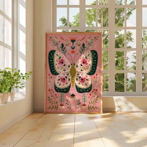 May include: A framed pink art print featuring a stylized butterfly with floral and leaf details. The butterfly has cream-colored wings with dark green and pink accents. The artwork is displayed in a room with a window and plants.