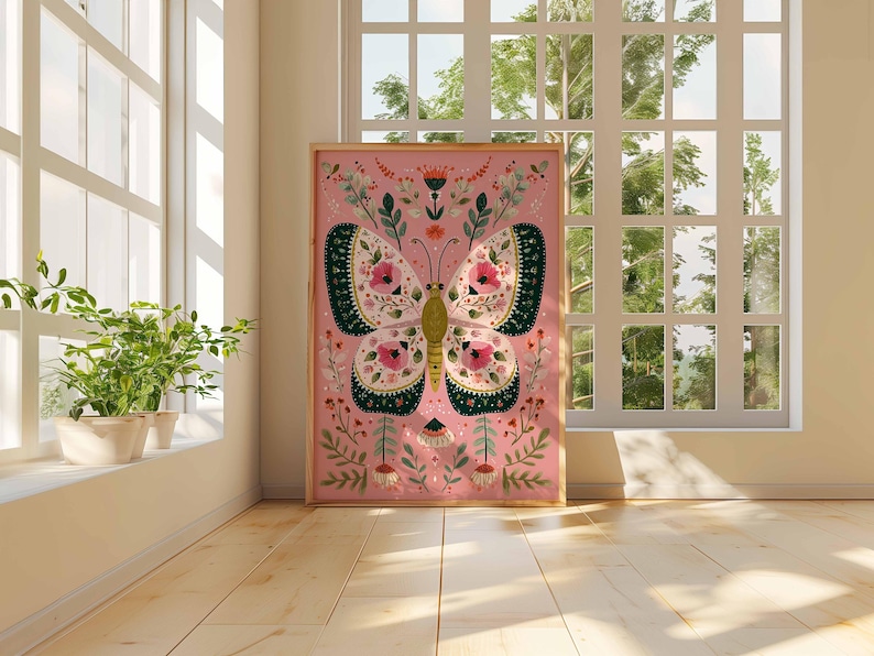 May include: A framed pink art print featuring a stylized butterfly with floral and leaf details. The butterfly has cream-colored wings with dark green and pink accents. The artwork is displayed in a room with a window and plants.