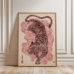 May include: Framed art print depicting a tiger in a descending pose, illustrated with dark red lines. The artwork is set against a cream backdrop with pink cloud patterns. Includes Japanese text and a red seal. The frame is a light brown colour.