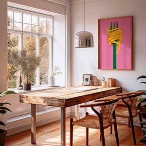 Op de afbeelding: Een eetkamer met een houten tafel, metalen poten en twee stoelen met vlechtwerk. Een ingelijst kunstwerk aan de muur toont een groene vork met gele pasta op een roze achtergrond. Een groot raam biedt uitzicht op bomen.